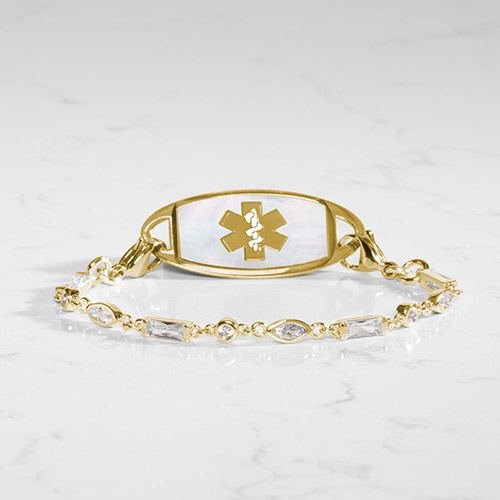 Gold-plated medical ID bracelet featuring a white mother-of-pearl tag with gold medical symbol and a crystal-accented link chain, displayed on a white marble background.