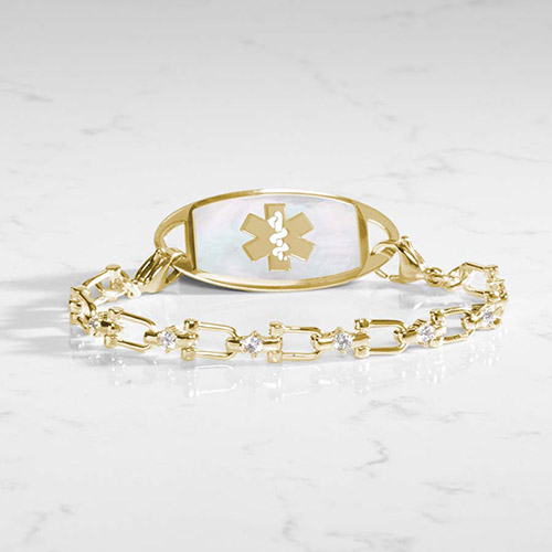 Elegant gold medical alert bracelet with mother-of-pearl ID tag and clear CZ-accented chain links, styled on a marble background.