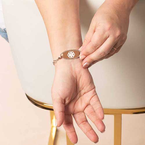 Woman displaying the front of the Diana medical ID bracelet in rose gold, highlighting the caduceus symbol and sparkling crystal frame on the tag.