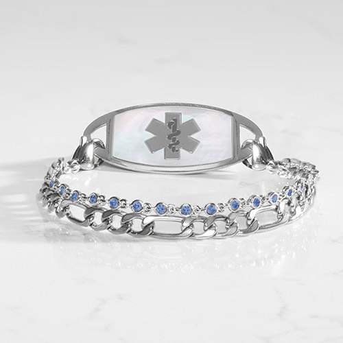 Silver women’s medical ID bracelet with white mother-of-pearl inlay tag and dual-chain design featuring blue crystal accents, displayed on a marble surface.