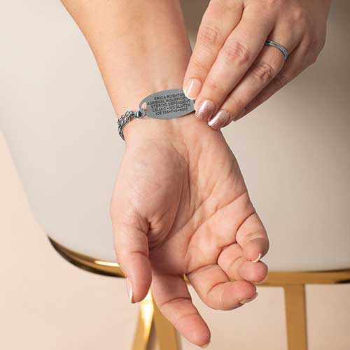 Close-up of a woman’s wrist wearing a custom-engraved silver medical alert bracelet with blue crystal details and double-chain style.