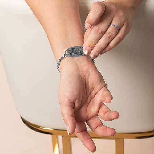 Close-up of a person’s wrist with a silver medical ID tag engraved with emergency information, held in place by a two-strand silver and lavender chain bracelet.