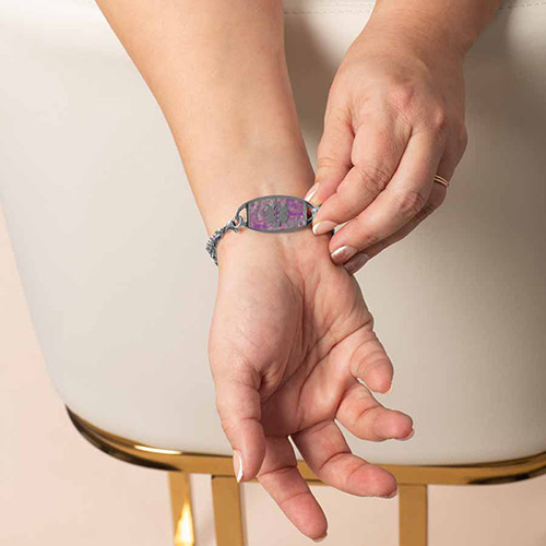 Close-up of a medical ID bracelet on a wrist, showing the front of the silver tag with a lavender and violet patterned inlay featuring a white medical caduceus symbol.