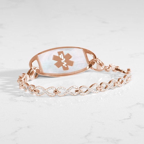 Rose gold medical ID bracelet with shimmering infinity links and a mother-of-pearl tag on a white surface.