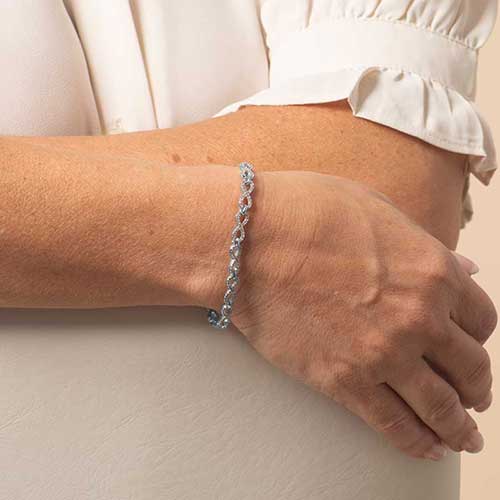 “Silver chain medical ID bracelet with sparkling infinity-style links worn on a woman’s wrist, shown against a short-sleeve cream blouse.”