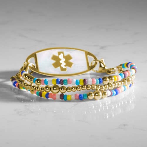 Gold tone beaded bracelet with colorful seed beads and oval medical ID tag displayed on a light stone surface.