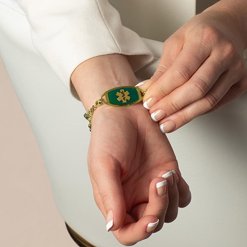 Laurel Medical Alert Bracelet with emerald and blue-green crystals, accented with gold, shown on wrist with Lauren’s Hope medical ID tag