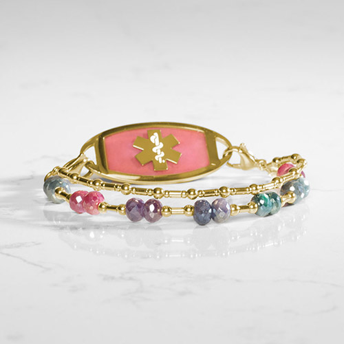 Medical ID bracelet with a gold pink-inlay tag and double-strand multicolor beaded band, displayed on a marble surface.