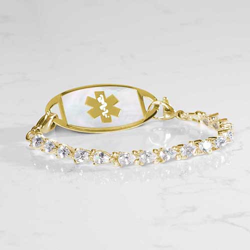 Gold medical alert bracelet with pear-shaped cubic zirconia stones and white mother-of-pearl ID tag engraved with gold medical symbol, styled on marble surface.