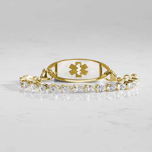 Gold medical alert bracelet with pear-shaped cubic zirconia stones and white mother-of-pearl ID tag engraved with gold medical symbol, styled on marble surface.