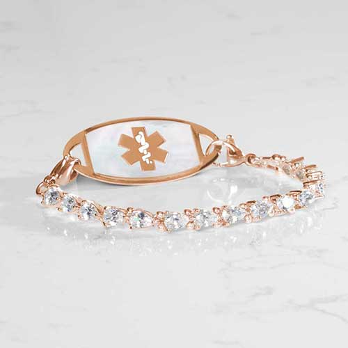 14k rose gold-plated women’s medical ID bracelet with pear-cut cubic zirconia stones and a white mother-of-pearl ID tag featuring the medical caduceus symbol, displayed on a marble surface.