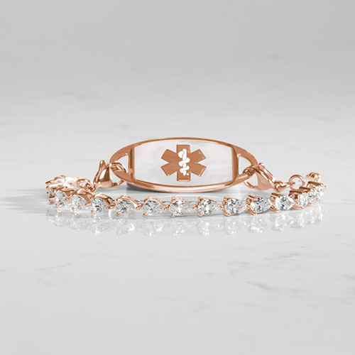 14k rose gold-plated women’s medical ID bracelet with pear-cut cubic zirconia stones and a white mother-of-pearl ID tag featuring the medical caduceus symbol, displayed on a marble surface.