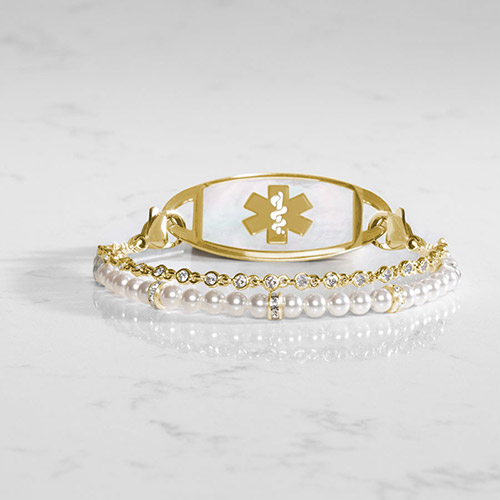 Gold medical ID bracelet with a mother-of-pearl tag and double-strand design featuring white pearls and bezel-set crystals, displayed on a marble surface.