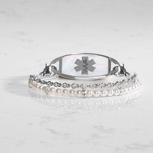 Silver medical ID bracelet with a mother-of-pearl tag and dual-strand design featuring white pearls and bezel-set crystals, shown on a white marble surface.