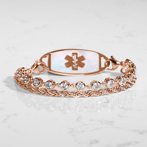 Rose gold medical ID bracelet resting on a marble surface, featuring a twisted chain and bezel-set CZ strand attached to a mother-of-pearl inlay ID tag with a rose gold caduceus symbol.