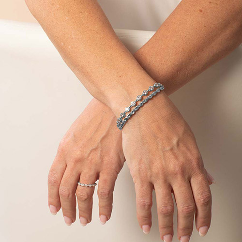 Woman crossing her hands to display the front of the Arden Silver bracelet on her left wrist, with two stacked strands—one of clear crystals, one of twisted chain—paired with a matching crystal-accented ring.