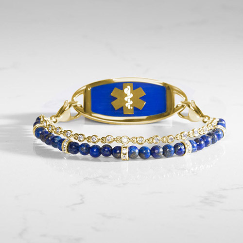 Women's gold medical ID bracelet with blue lapis beads and clear crystal accents, featuring a navy medical alert tag with gold caduceus symbol, displayed on a marble surface.