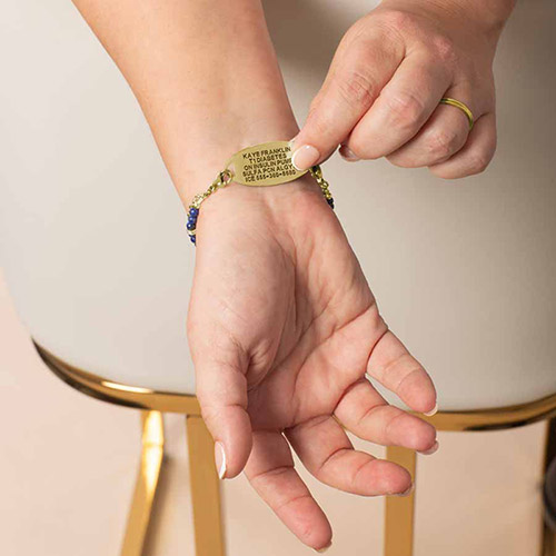 Close-up of a woman's hand adjusting a gold medical ID bracelet with an engraved nameplate, featuring blue and gold beads, designed for stylish diabetes alert and emergency medical information.