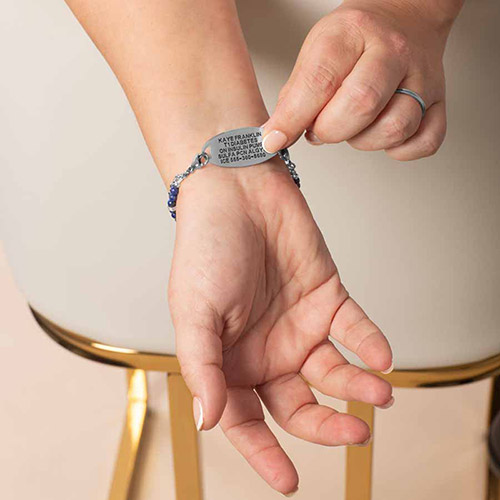 Close-up of Charlotte Blue women's medical ID bracelet in silver, showing custom-engraved tag with medical alert information being held open on wrist.