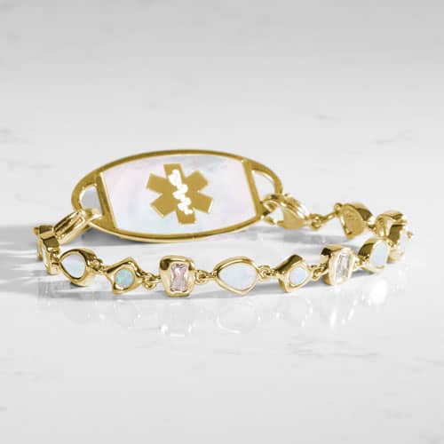 Gold-tone medical ID bracelet with a mother-of-pearl ID tag and alternating opalescent and light pink stones on a marble surface.