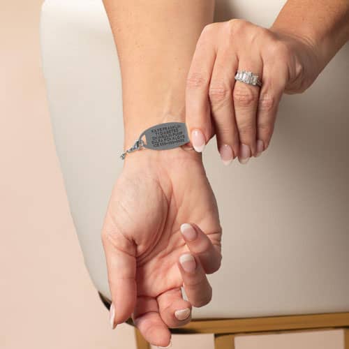 Model lifts the blue medical ID tag to display the engraved medical information on the back of the blue beaded bracelet.