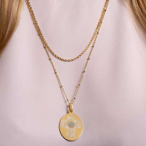 Woman in pink top wearing gold tone oval pendent with detailed cross patterned and layered necklace chains.